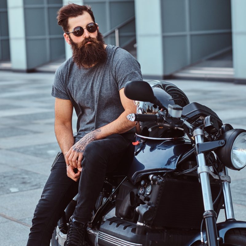 brutal-bearded-male-in-a-gray-t-shirt-and-black-pants-sitting-on-his-custom-made-retro-motorcycle.jpg